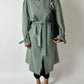 Person wearing a long green coat with a belt, holding a black bag on a white background
