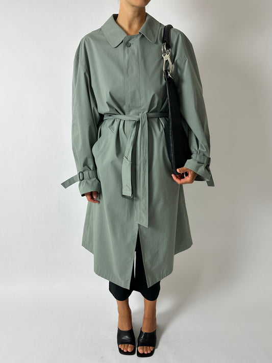 Person wearing a long green coat with a belt, holding a black bag on a white background