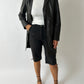 Person wearing a black coat, white shirt, black shorts, and black strappy shoes on a white background