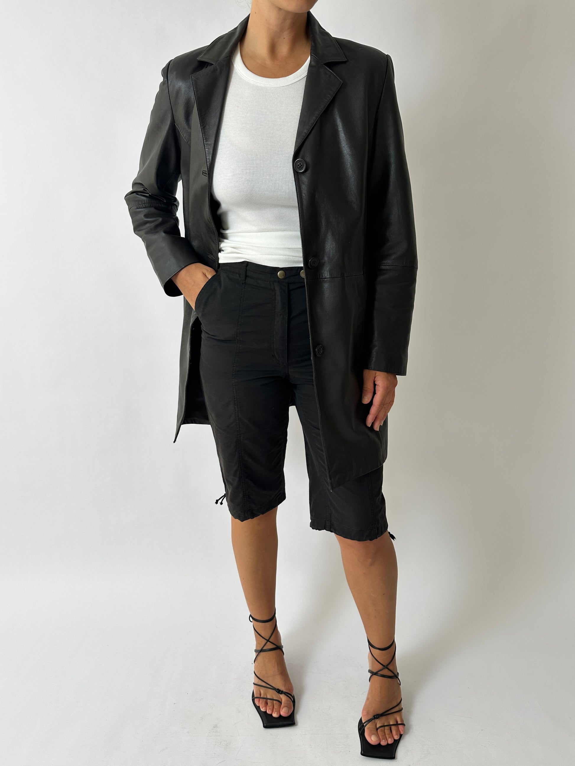 Person wearing a black coat, white shirt, black shorts, and black strappy shoes on a white background