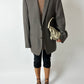 Person wearing a gray blazer over a patterned top with black pants and high heels, holding a beige handbag on a white background.