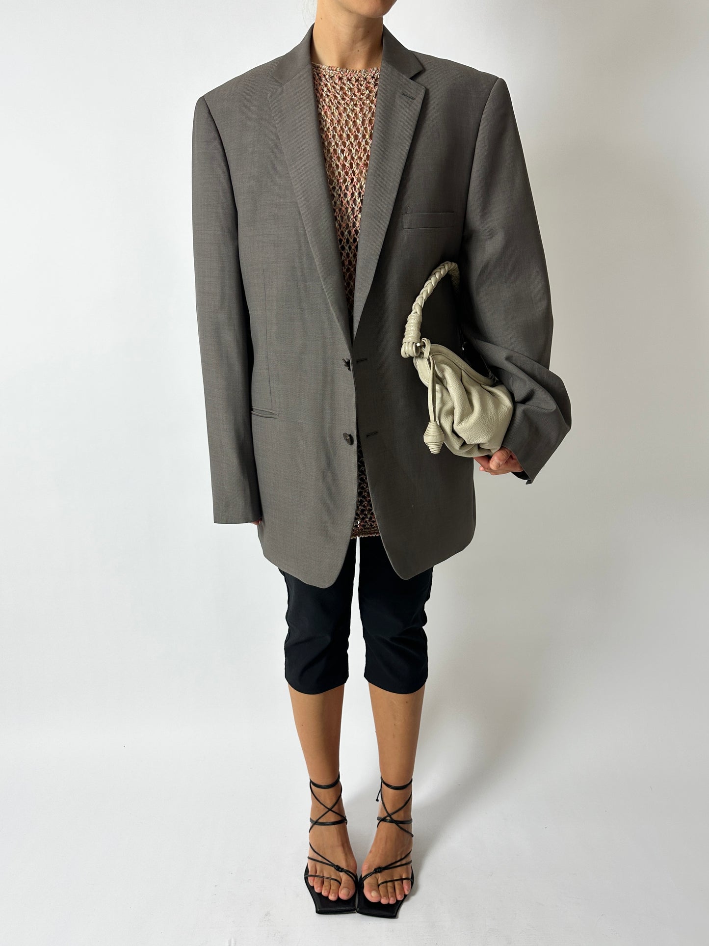 Person wearing a gray blazer over a patterned top with black pants and high heels, holding a beige handbag on a white background.