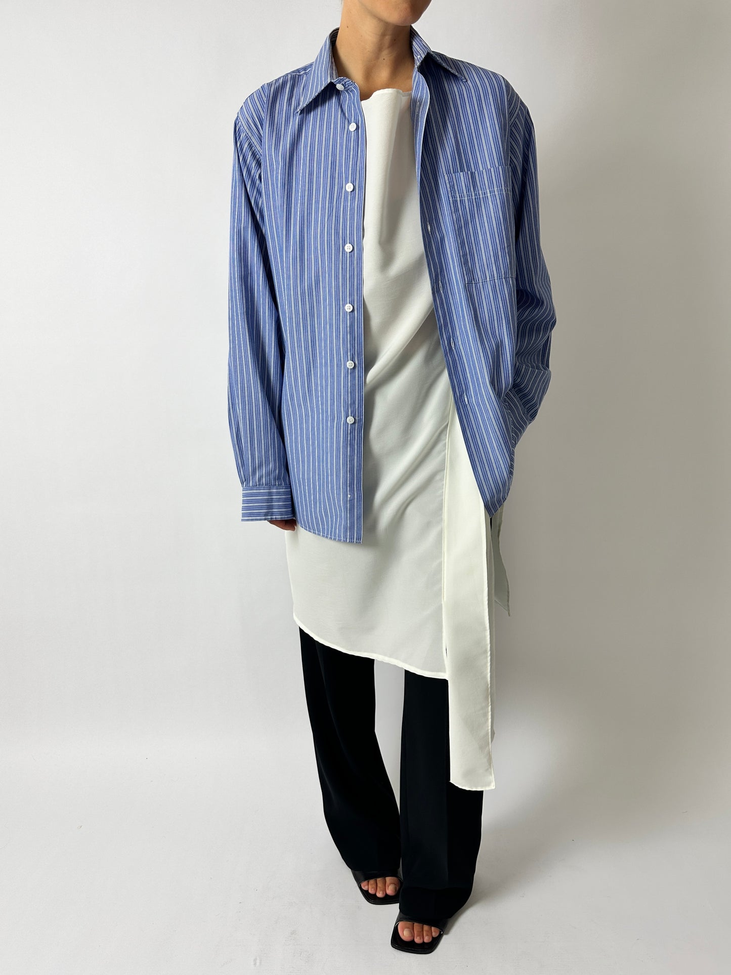 Person wearing a blue striped shirt over a white top with black pants on a plain background