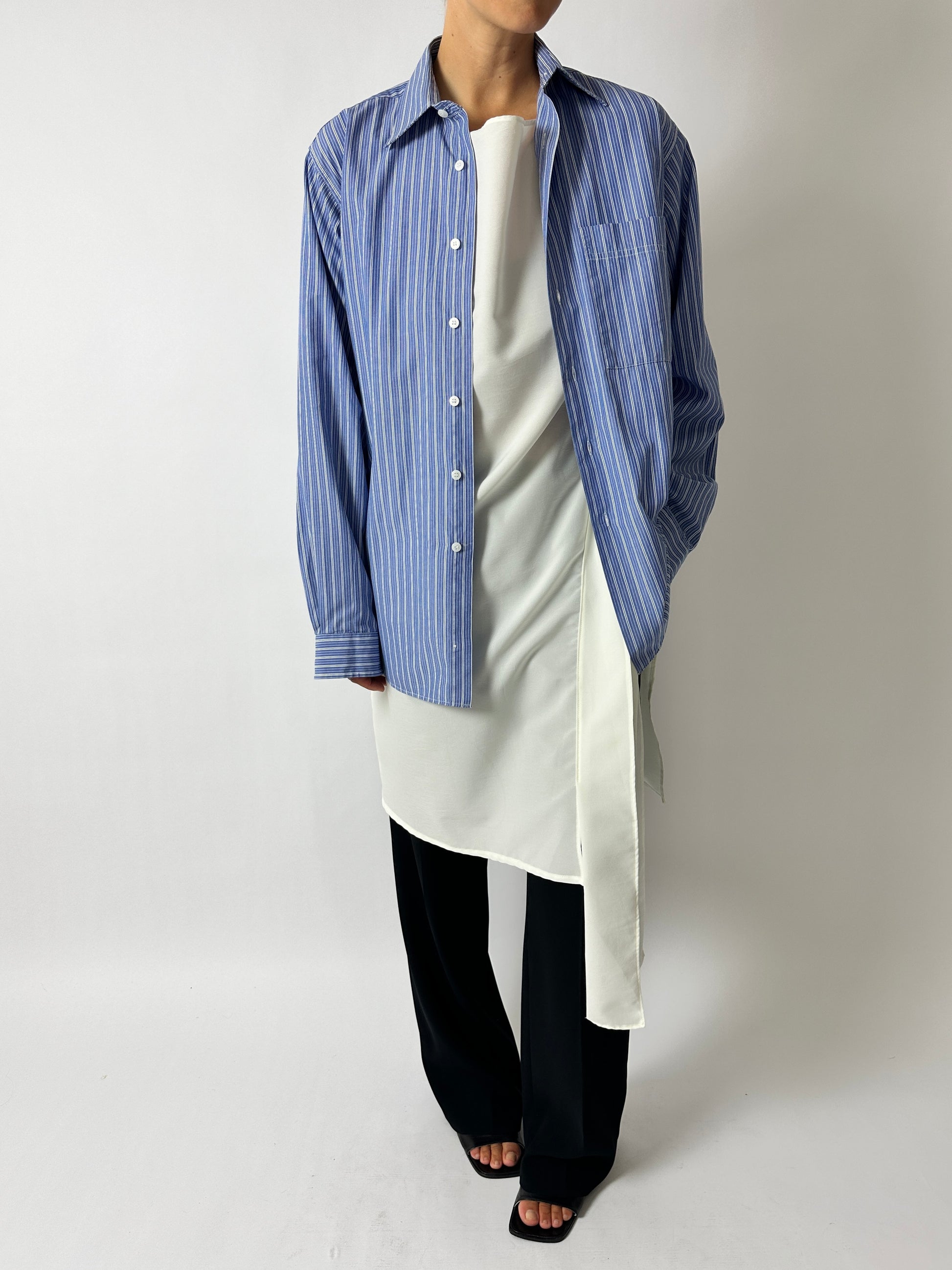Person wearing a blue striped shirt over a white top with black pants on a plain background