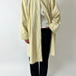 Person wearing a beige long coat over a white shirt and black pants on a plain background