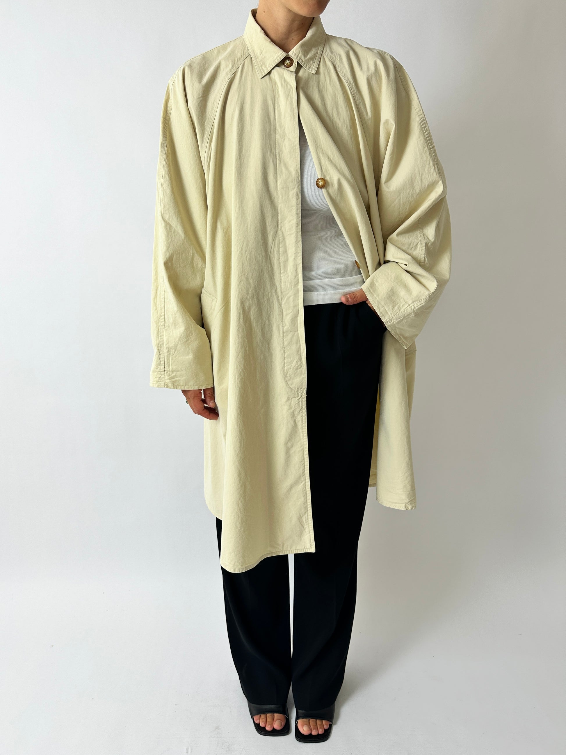Person wearing a beige long coat over a white shirt and black pants on a plain background