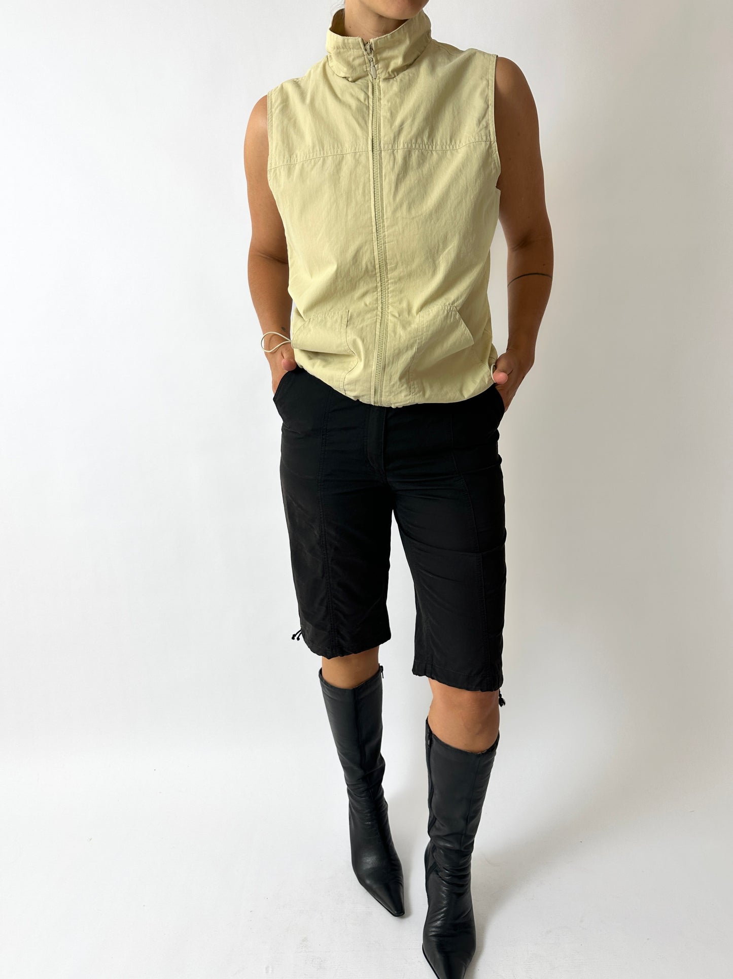 Person wearing a light yellow sleeveless jacket, black pants, and black boots on a white background