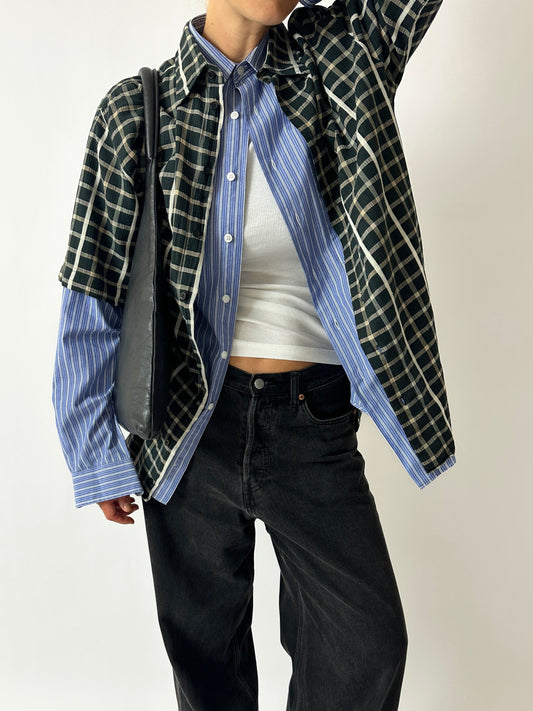 Person wearing a layered outfit with a plaid shirt, striped shirt, and black pants on a plain background