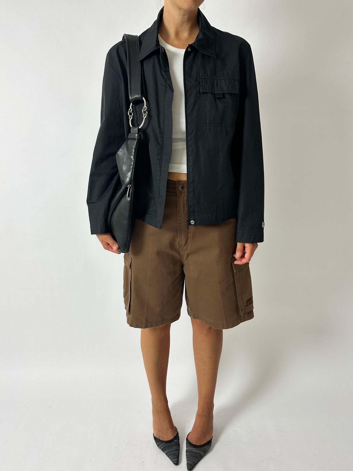 Person wearing a black jacket, white shirt, brown shorts, and black shoes on a white background