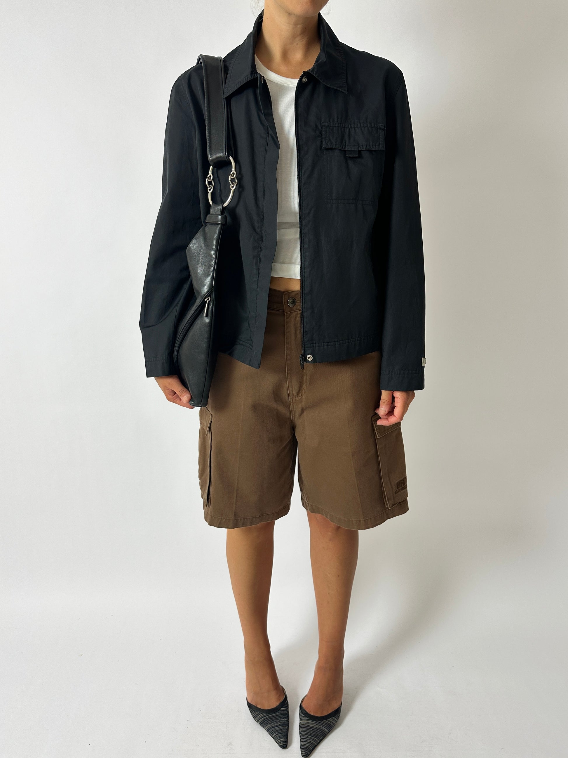 Person wearing a black jacket, white shirt, brown shorts, and black shoes on a white background