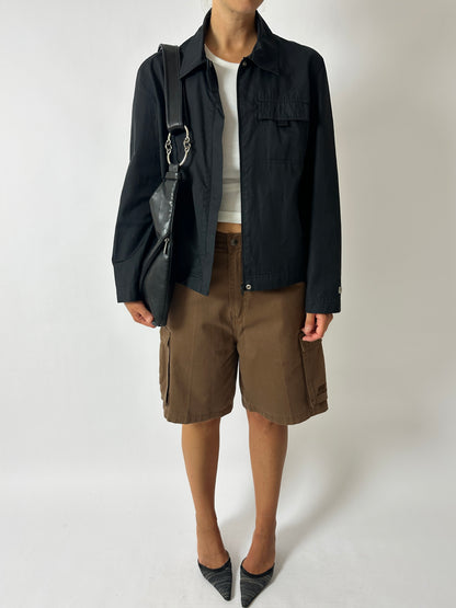 Person wearing a black jacket, white shirt, brown shorts, and black shoes on a white background